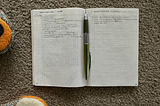 Photo of an open A6 notebook with handwritten lists and a mechanical pencil in the centre. The book is lying on carpet and there are glimpses of a little girl’s slippers nearby