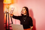 A perfectly groomed, white woman poses with her phone in front of a pink wall. Her trendy outfit is surely fulfilling her fans dreams while showing off her influencer/content creator chops.