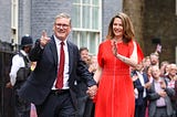 The image shows Keir Starmer, the new Prime Minister of the United Kingdom, walking hand-in-hand with his wife, Lady Starmer. Both are smiling and waving to a crowd gathered around them. Keir Starmer is dressed in a dark suit with a red tie, and Lady Starmer is wearing a bright red dress. A police officer and several people holding British flags can be seen in the background.