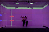 To dancers perform in a room with purple lighting, responding to projected lines and shapes on the walls. The digital visuals from a grid-like score that guides their improvised movements.