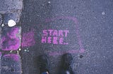 Start Here written on pavement with two feet in black shoes at the starting line.