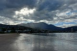 view of Hobart city from Long Beach, Sandy Bay. it is almost night, the sun is setting, but the sky is cloudy, with sun coming through in patches. The water is calm and glassy. The hills are dark except for one in the middleground, where the author’s home is situated.