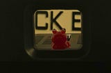A small red toy frog with large round eyes sits centered inside a narrow rectangular opening, with blurred black letters in the background. The framing creates a tunnel-like view that draws attention to the bright, playful figure.