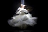 A fotografia captura mais de um momento da dança uma mulher que está usando um vestido branco de tamanho midi. A imagem está mexida. O vestido se mexe, o rosto, e os braços, como se tivesse sido capturada em um momento de rodopio.