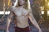 A scene from the TV series “The Vampire Lestat.” It shows actor Sam Reid as Lestat, standing on a concert stage during a performance. He has on no shirt and is wearing tight pants. A drum set is behind him.