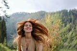 A woman with long red hair smiles as her hair flows around her. She is wearing a tan v-neck dress and standing in front of the woods.