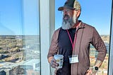 The author, a bearded man wearing glasses, a baseball cap, and a dark long-sleeve shirt, stands in front of a tall glass window overlooking a cityscape. He holds a coffee cup in one hand and wears a visible name badge that reads “Andrew.” A pink lanyard is around his neck, and he has visible tattoos on his forearm and a smartwatch on his wrist. The background shows a clear blue sky, buildings, and trees.