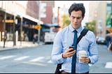 Half-portrait of businessman walking down street wearing blue button down shirt and tie, with messenger bag draped on shoulder, frowning at cell phone in one hand and carrying take-out coffee in the other hand.