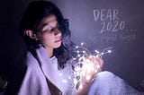 Sitting with fairy lights that are in a ball, wearing a scarf. Labeled, “Dear 2020…my deepest thoughts”.