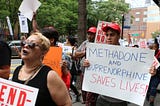 Protesters stand with signs, large white sign says “Methadone and Buprenorphine Saves Lives”