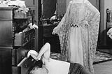 A man and woman in a messy room, and the man is trying to find something underneath the dresser.