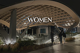 In the background: a looming wooden mesh trellis, an architectural sculpture situated in an urban landscape. Beneath it, three women stand talking and gesturing excitedly. In the foreground: white text that reads: WOMEN. Beside the text, a hand drawn, squiggly arrow points towards the women in the background image.