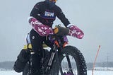 Patricia George, the article author, riding on a Why Cycles Big Iron fat tire bike in the snow with a Team PHenomenal Hope jacket and purple and pink camoflage pogies. The bike has all her camping gear on it as well.