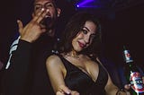 Man and woman in a party scene. Man grinning, making a hand sign, woman in a low cut dress, smiling while holding a beer.