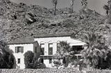 Black and white photograph of The Willows in Palm Springs. The Mediterranean-style villa sits at the base of a rocky hillside with the San Jacinto Mountains rising behind it. The white stucco exterior features arched windows, a tiled roof, and a small balcony, with palm trees and desert landscaping surrounding the residence.
