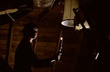 A screencap from The Exorcist (1973) in which Chris MacNeil searches a cluttered attic for signs of a disturbance. She’s lit only by a single candle held in her outstretched hand.