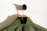 Photograph of the back of an army green jacket, hanging from a hanger. The collar has been sliced in half, and a black leather triangular patch has been sewn in to create a ‘collar gap’.