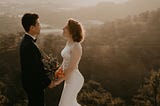 Groom and bride stand holding hands with a bouquet at the top of a mountain, overlooking a pretty vista.