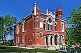 A photo of the ornate Jewish temple, Temple Aaron, which was founded in 1883 in Trinidad, Colorado. It’s red brick, with two turrets and a big clock at the front. Large stone pillars frame the entrance.