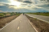 A paved bike path at sunrise with a lone bike rider in the distance.