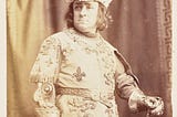 Black and white photo. Man in rich 15th-century dress. Belted tunic decorated with fleur de lys and lions, hat and gauntlets.