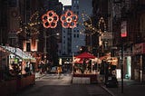 A street shot of a Little Italy in the States. Decorative streetlights in the shape of red flowers create an archway over the street. There are small food stalls slipping out of the sideway and onto the narrow city street.