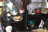 A close up shot of two small black side tables with household items such as saucepans, a coffee maker, a juicer, and iron, some baking ware. All this is on a front porch, ready to give away.