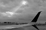 An airplane wing is in the center. The sky has thick clouds with a window where a column of sunlight comes through.