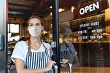 Photo description: Young woman in striped apron and mask stands at the door to a bakery