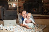 father and son sitting on the floor in front of a fire with a laptop open