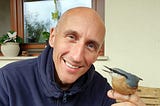 The author, József Manhertz, looks into the camera with a smile. He is bald and has blue eyes. A Eurasian nuthatch sits on the index finger of his left palm.