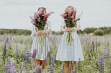 Two sisters in knee-length dresses in a lilac field holding colorful bouquets of flowers outstretched in front of themselves, hiding their faces.