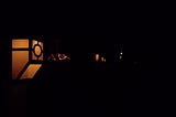 A nighttime street photo of mostly darkness; but a row of fence-post amber lights recedes from left to right into the distance. Very small at the top of the photo is the moon, a tiny bright dot; and far away in the right side of the photo are two more tiny bright pale dots, streetlights.