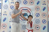 Father and Son in front of a banner after a Taekwondo competition. The son has a red belt and it wearing a Taekwondo uniform