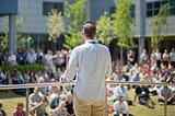 A man speaks to a crowd, who are seated on the ground in front of him