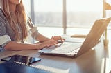 Woman blogging in spacious office using computer on her workplace. Female employee sitting, smiling, looking at camera.