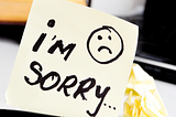 This image shows a yellow sticky note with the handwritten message “I’m Sorry…” accompanied by a sad face drawing. The note is placed on a desk near a keyboard and some crumpled paper.