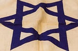 A hand-sewn flag, feature a blue Star of David at the center and two horizontal blue cloth stripes near the top and bottom.