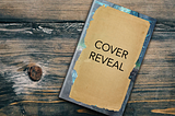 A book sitting on a wooden table. The cover of the book is covered with a piece of brown paper featuring the words “Cover Reveal.” The edges of the book cover can be seen around the edges of the paper, and the cover color is blue-green.