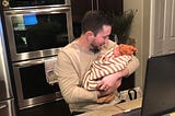 Brett holding baby Ezra in the kitchen while working on a laptop. Brett is looking down and smiling at Ezra. A Platform 9 and 3/4 magnet can be spotted in the background.