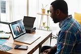 A black man staring into a laptop screen. The laptop is on a wooden desk. A woman’s face is on the laptop screen. Their posture and set-up models a virtual interview.