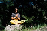 A young, dark haired woman in grey crop top and yellow leggings sitting cross legged on a rock, meditating with her palms together. She is surrounded by greenery.