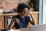 Young woman of color presses a finger to the bridge of her nose in clear frustration as she sits in front of a laptop.