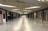 An empty corridor in Melbourne’s Tullamarine Airport during one of the longest Covid-19 lockdowns in the World. Diagonal strips of light are stepped along the low ceiling, reflecting off the sheen of the off-white tiled floors. All of the shops are closed.