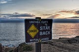 Signage upon a body of water that reads “Unstable Cliffs Stay Back”