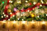 candles in foreground that spell out “peace” with the twinkling of christmas tree lights in background