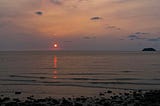 Sunset over the sea. Small orange sun is almost at the horizon, reflected in the water. A small island is on the right side of the photo on the horizon. A rocky shore is in the foreground.