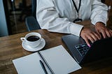 woman typing with a cup of coffee to the side and an empty journal