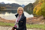 Susan faces the camera, she’s sat in front of a beautiful lake and is wearing a scarf and coat