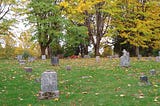 Photo of slanted cemetery with decaying old graves and autumnal leaves on the ground, trees in distance.